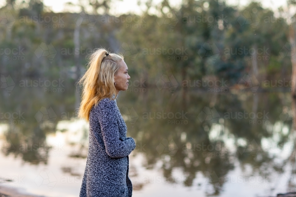aboriginal woman by herself in tranquil setting with reflections of trees on water - Australian Stock Image