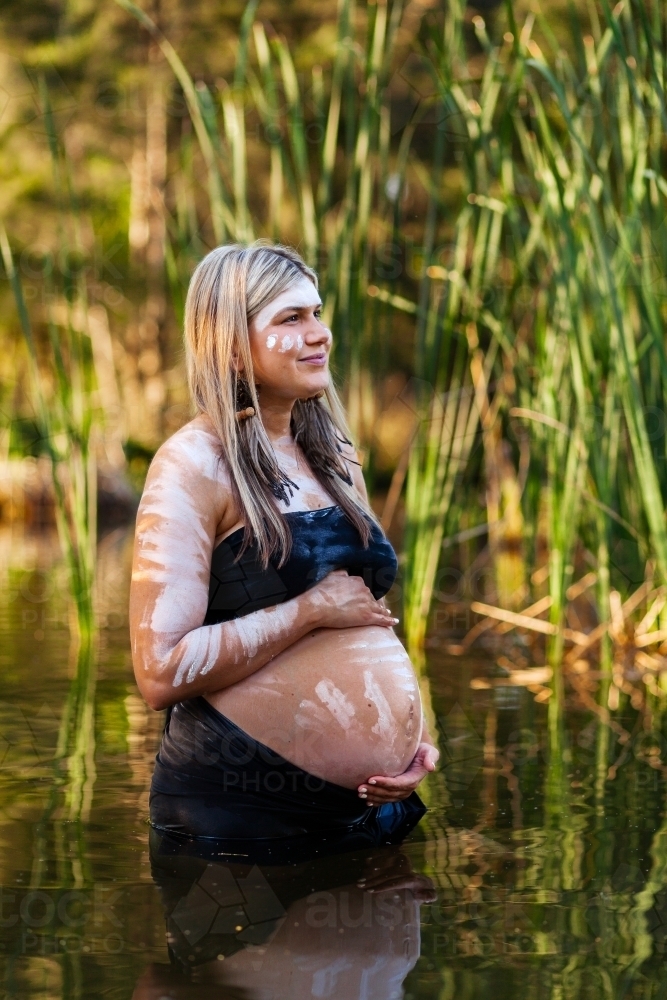 Aboriginal woman in traditional body paint standing among reeds in billabong - Australian Stock Image