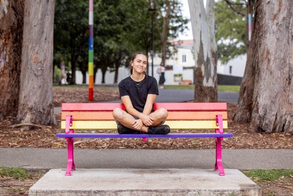 Aboriginal woman sitting cross-legged on a rainbow park bench - Australian Stock Image