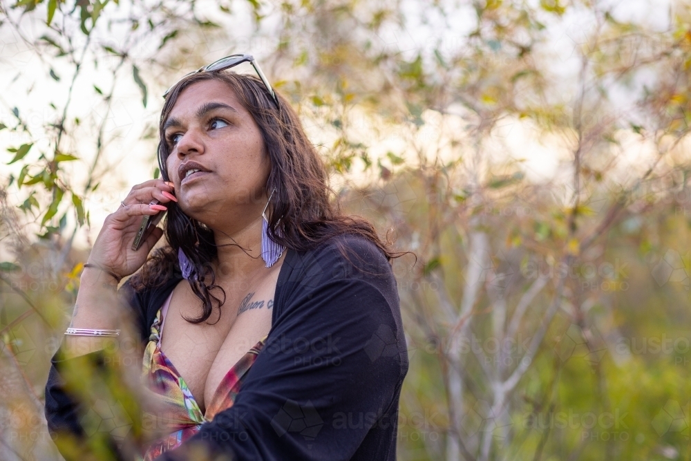 Aboriginal woman with sunglasses on head talking on mobile phone with bush in background - Australian Stock Image
