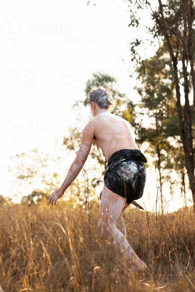 Aboriginal young man in traditional ochre body paint stalking kangaroo through grass in paddock - Australian Stock Image