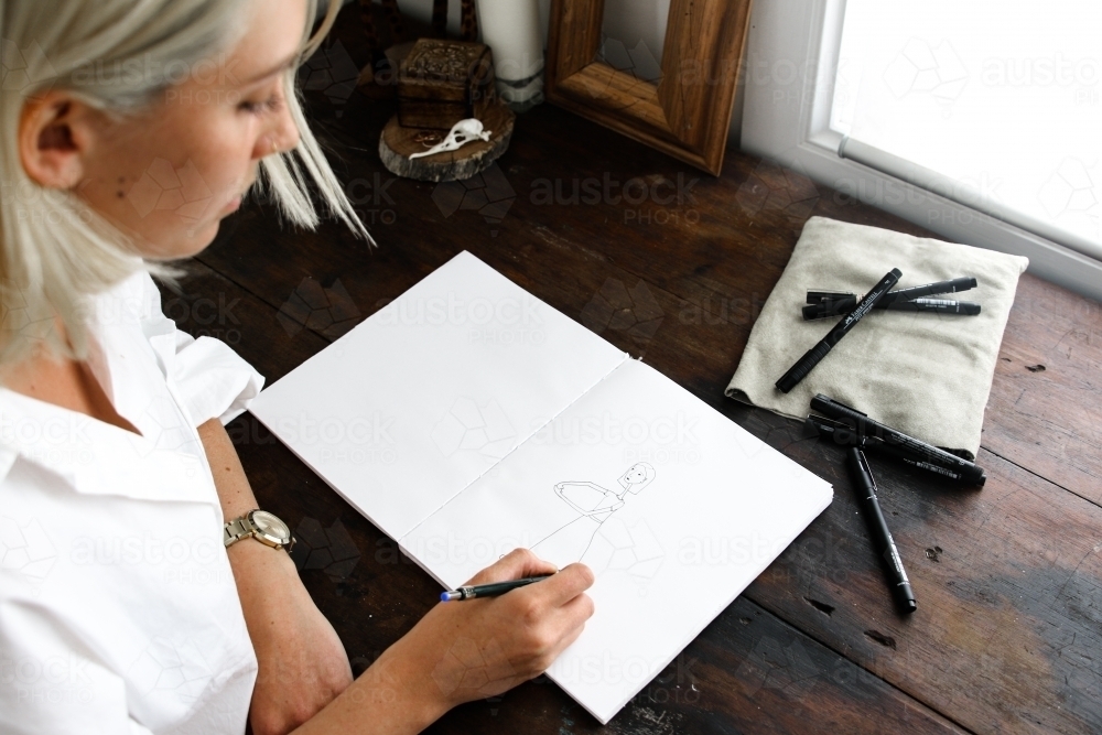 Above over the shoulder shot of young blonde woman drawing in sketch book with pens - Australian Stock Image