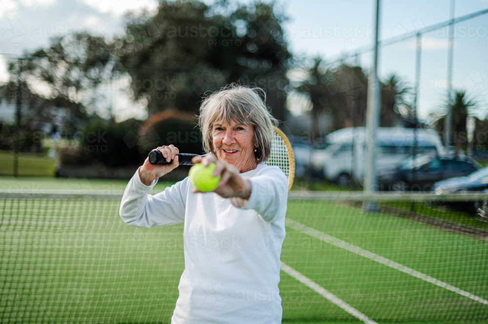 Active elderly woman enjoys tennis at an outdoor court, ready to serve with determination - Australian Stock Image