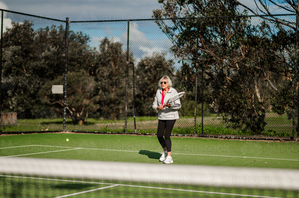 Active senior woman enjoying a game of tennis outdoors on a bright day, wearing sunglasses. - Australian Stock Image