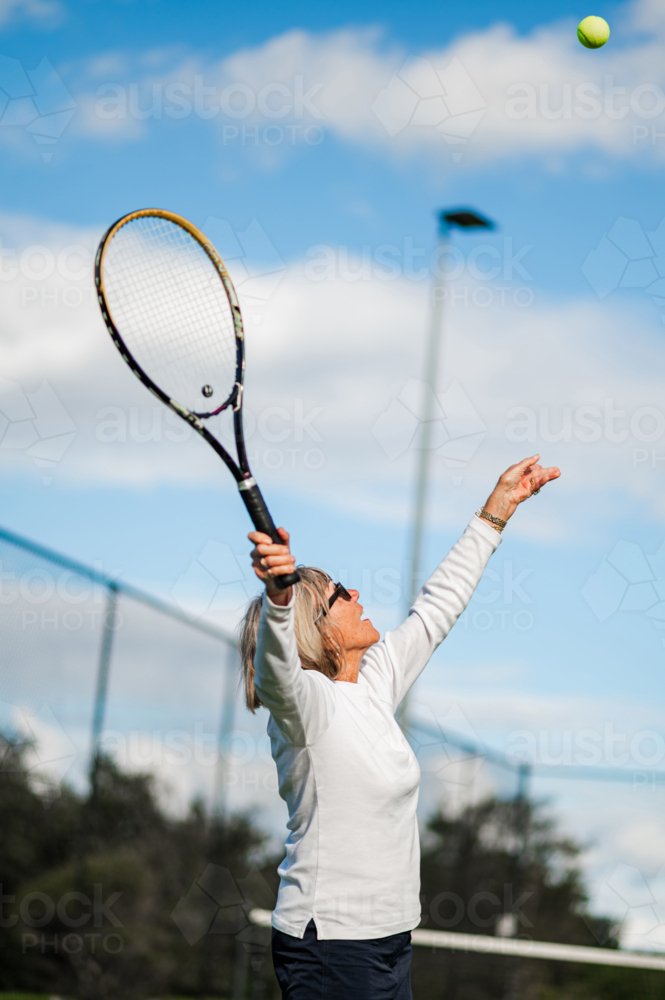 Active senior woman serves a tennis ball outdoors at a sunny tennis court - Australian Stock Image