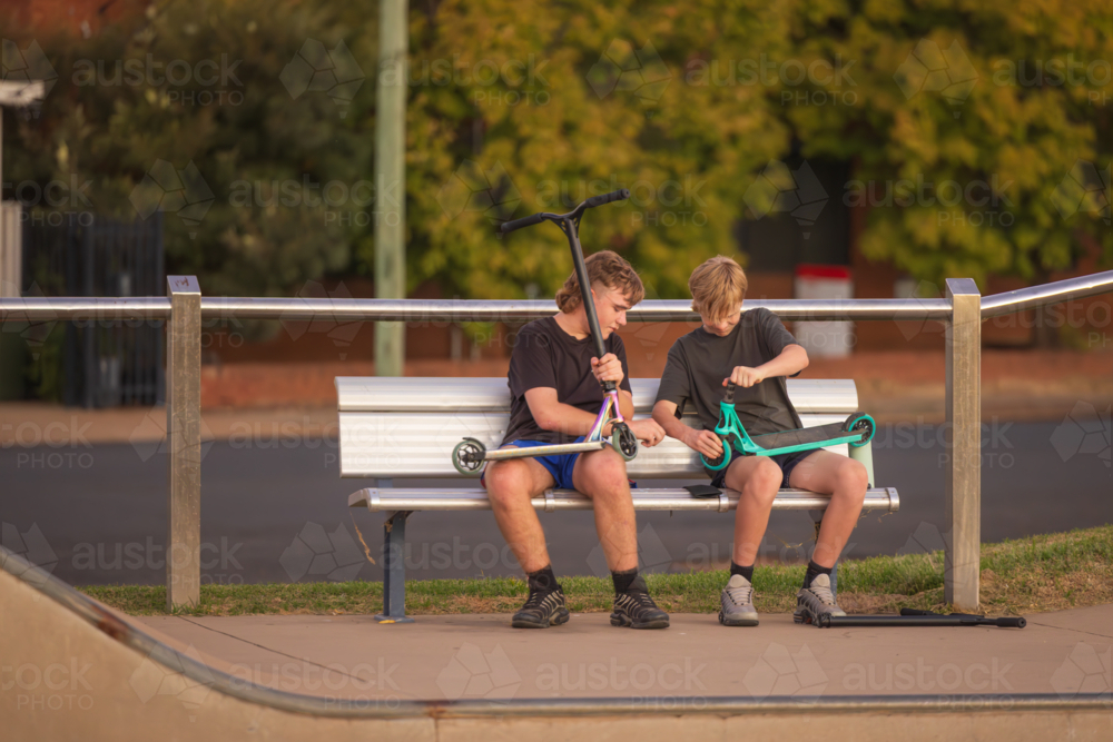 Adolescent teenage boys sitting on bench at skate park making repairs to scooters - Australian Stock Image