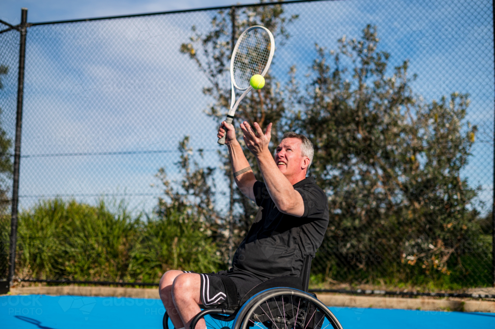 Adult male athlete in wheelchair prepares to serve tennis ball on bright day in outdoor setting - Australian Stock Image