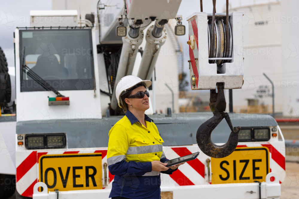 adult woman in reflective clothing holding a tablet on the worksite by oversize crane truck - Australian Stock Image