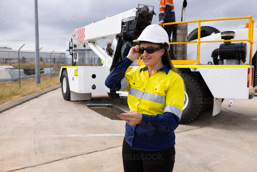 adult woman in reflective clothing holding a tablet on worksite by crane - Australian Stock Image