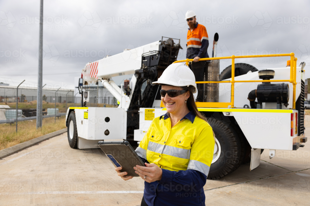 adult woman in reflective clothing holding a tablet on worksite by crane - Australian Stock Image