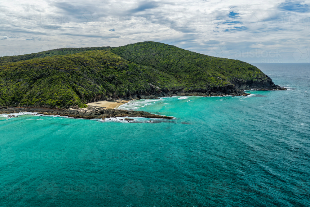 Aerial drone view of Sharkies Beach featuring a rugged rocky coastline and a tree covered mountain. - Australian Stock Image