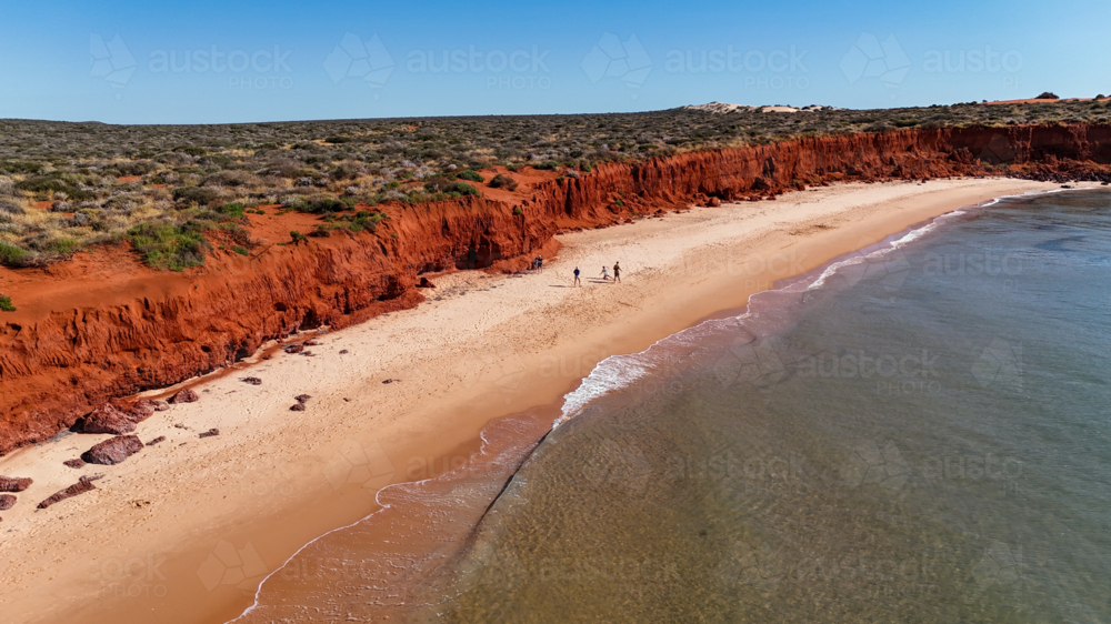 Aerial of family walking along beach where red cliffs meet white sand and water - Australian Stock Image