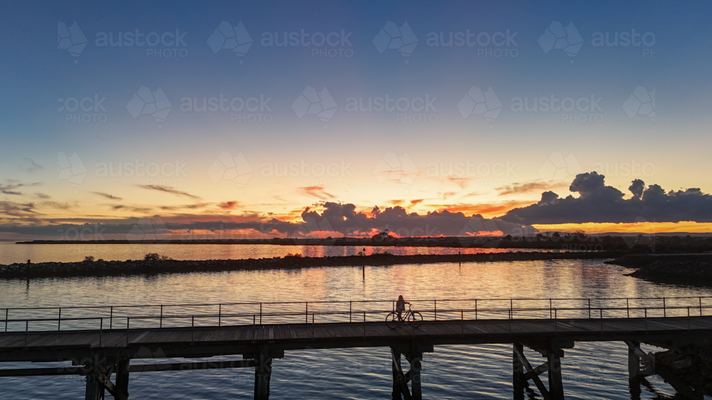Aerial of silhouette of person on bike riding along jetty at sunset - Australian Stock Image