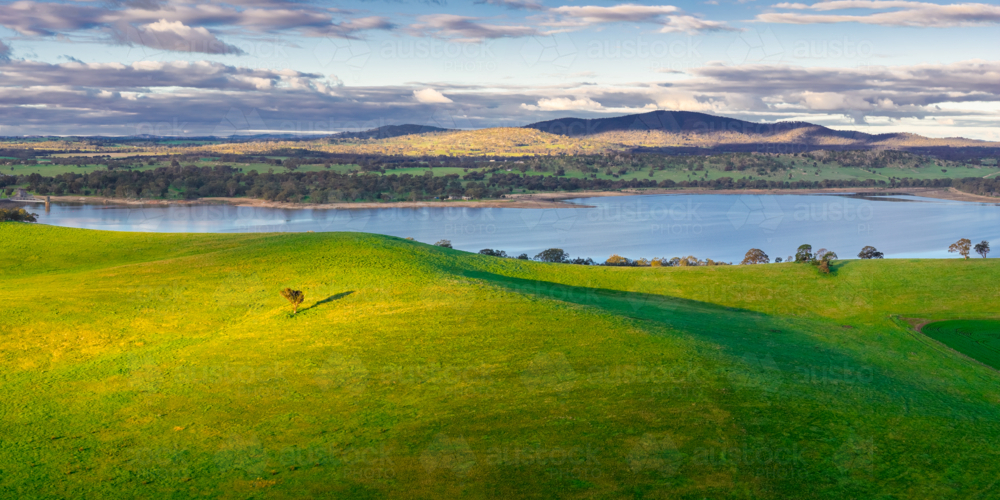Aerial panorama view of sunshine on lush green hills around an inland lake and distant hills - Australian Stock Image