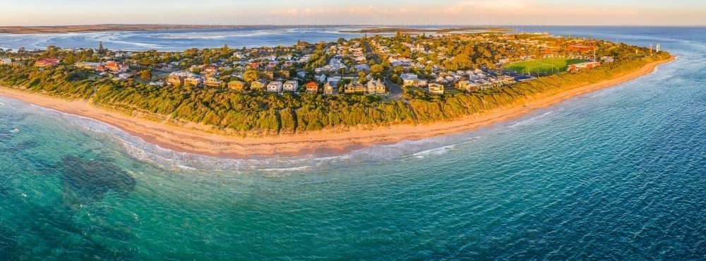 Aerial panoramic view of a sandy beach and coastal town stretching along a peninsula at sunset - Australian Stock Image