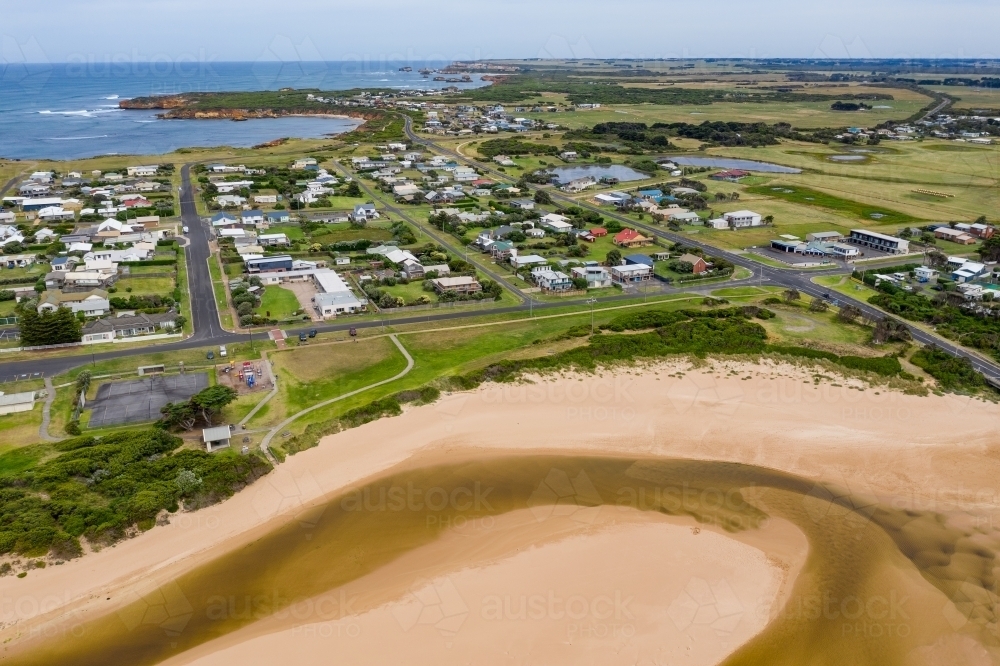 Aerial view a creek flowing over a sandy beach below a coastal town - Australian Stock Image