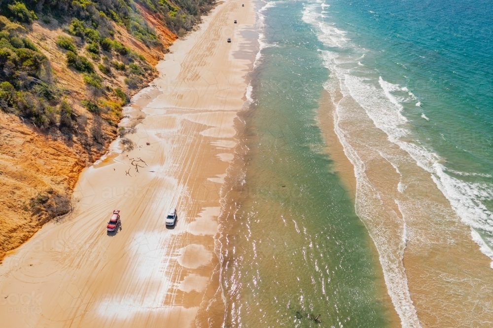 Aerial view of 4WD's on a narrow sandy beach between waves and high coloured cliffs - Australian Stock Image
