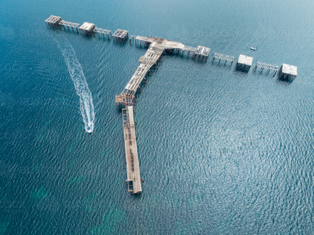 Aerial view of a boat speeding past an abandoned pier over a turquoise ocean - Australian Stock Image