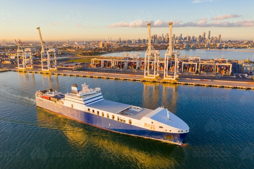 Aerial view of a cargo ship cruising past large gantry cranes on a dock - Australian Stock Image