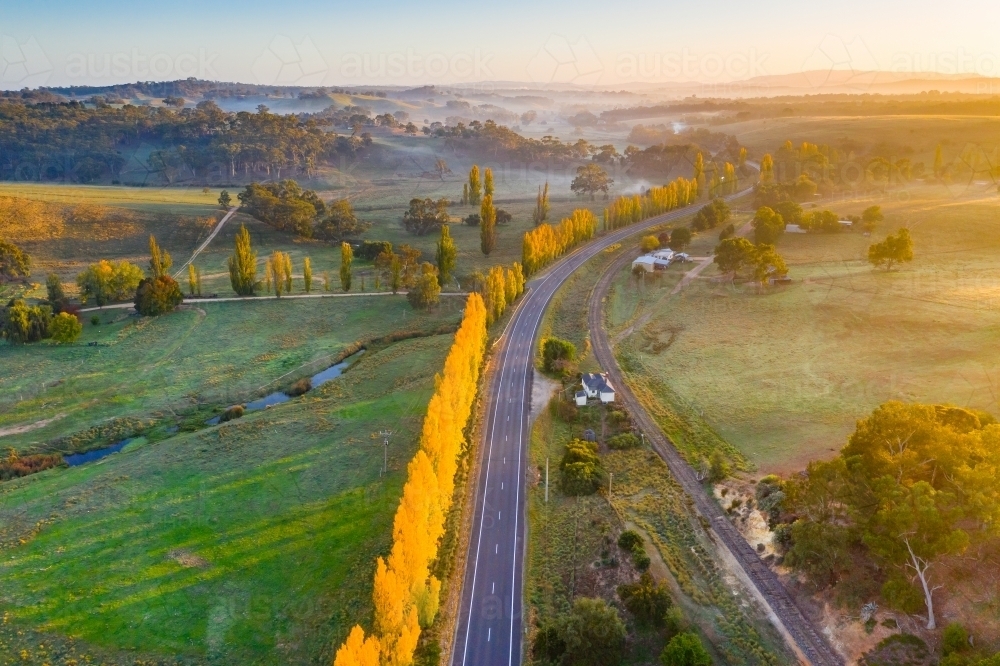 Aerial view of a country road winding through autumn trees at sunrise - Australian Stock Image