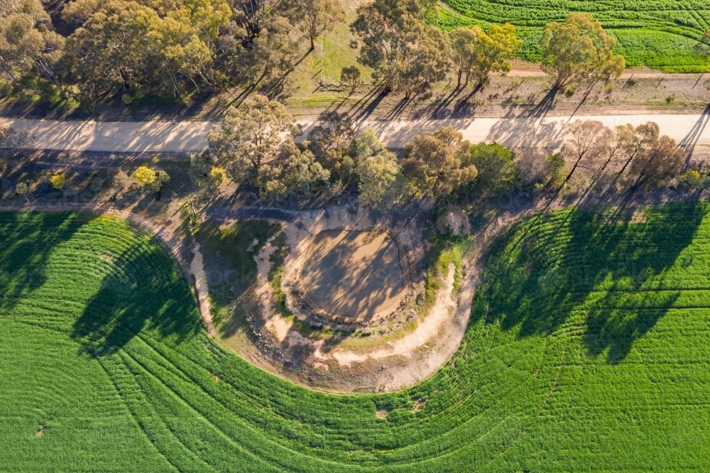 Aerial view of a dirt road and dam between green paddocks with overhanging gum trees - Australian Stock Image