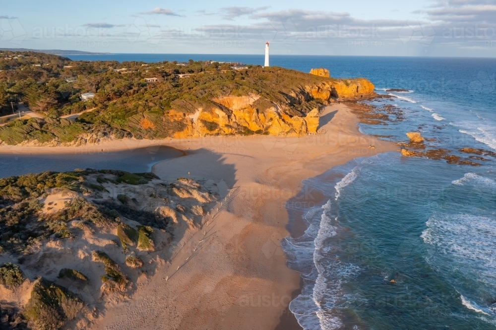 Aerial view of a lighthouse high on sea cliffs above a sandy beach in afternoon sunshine - Australian Stock Image