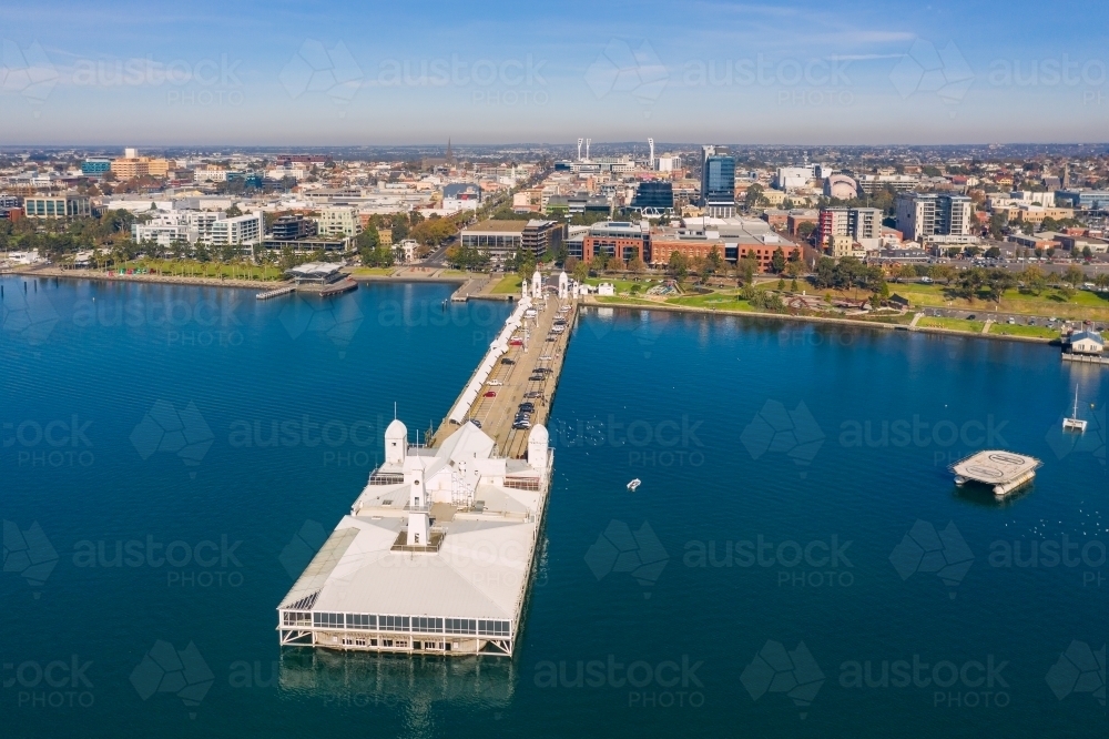 Aerial view of a long straight jetty with buildings on the end and a coastal city in the background - Australian Stock Image