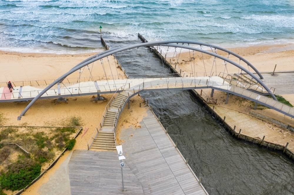 Aerial view of a modern arched walking bridge over a marina channel flowing out to sea. - Australian Stock Image