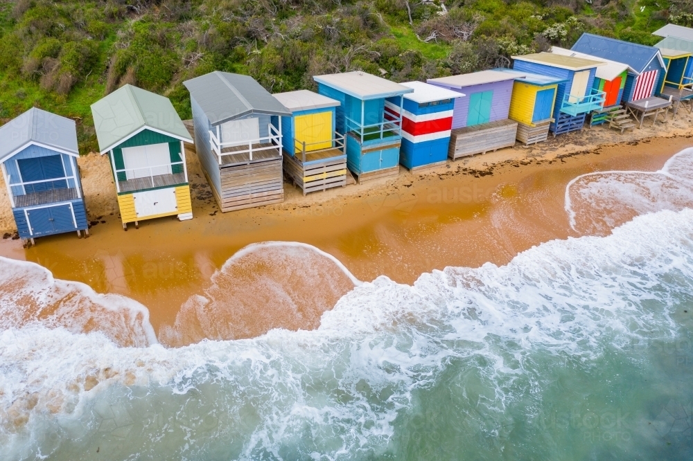 Aerial view of a row of colourful beach huts with waves washing up underneath - Australian Stock Image