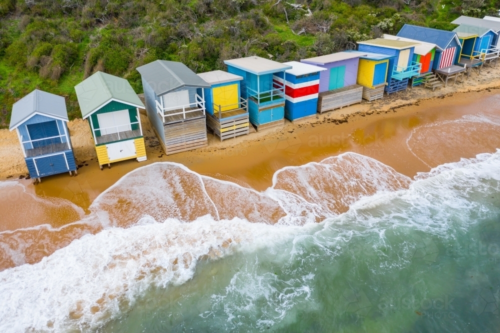 Aerial view of a row of colourful beach huts with waves washing up underneath - Australian Stock Image