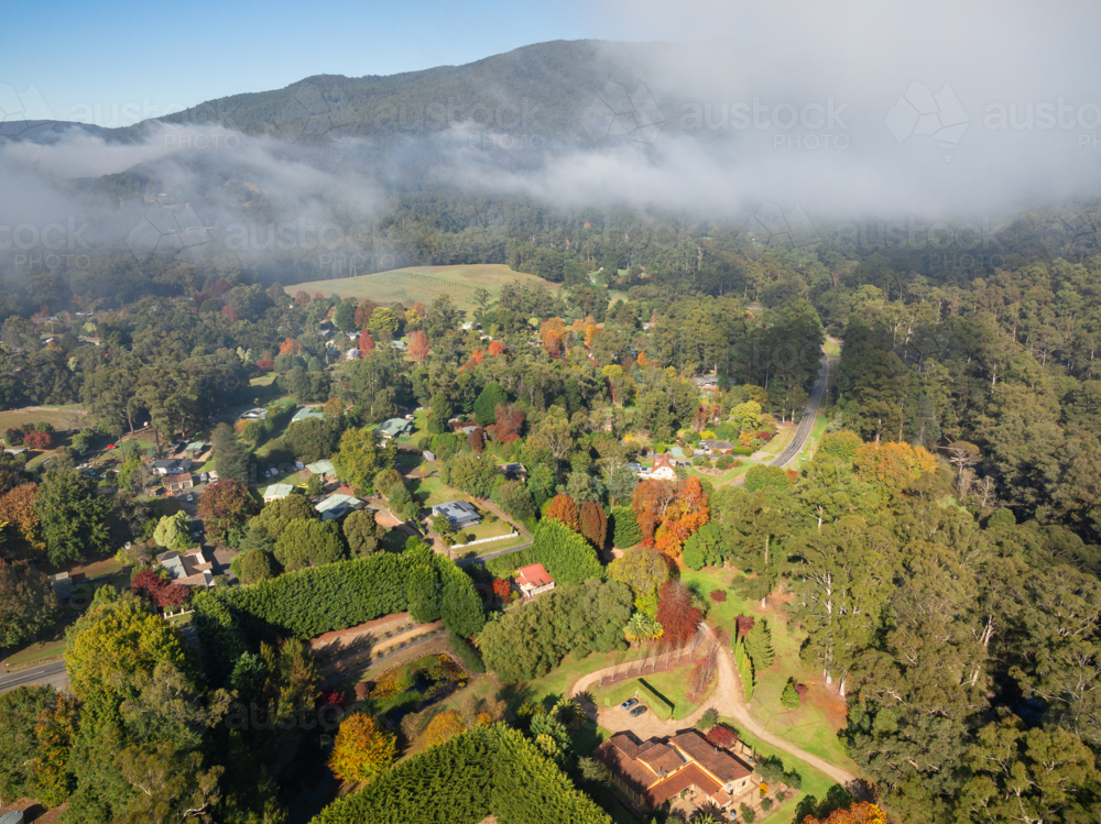 Aerial view of a rural town in a mountain valley with lingering clouds and colourful Autumn foliage - Australian Stock Image