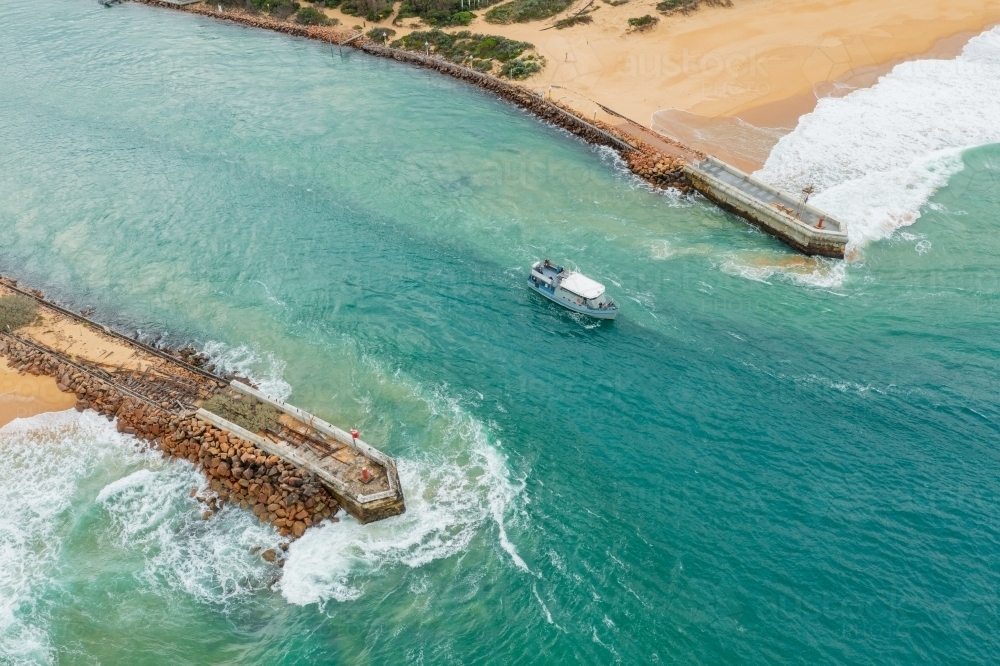 Aerial view of a small boat going out to sea between breakwaters through turbulent water - Australian Stock Image