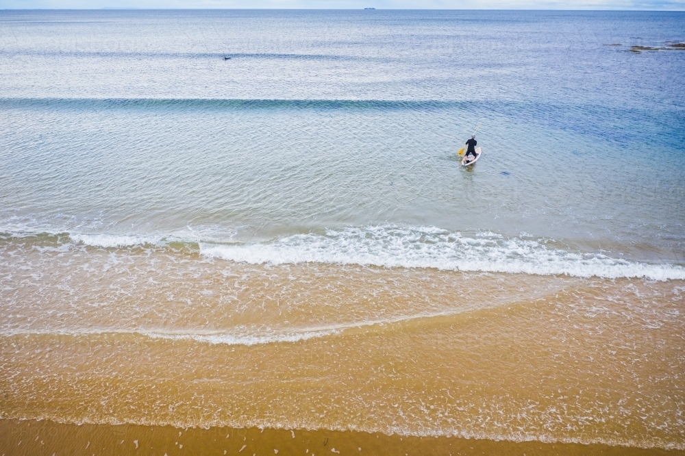 Aerial view of a stand up paddle boarder rowing out to sea over small waves - Australian Stock Image