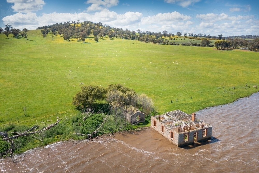 Aerial view of an abandoned historic farm house full of water on the edge of a lake below green hill - Australian Stock Image