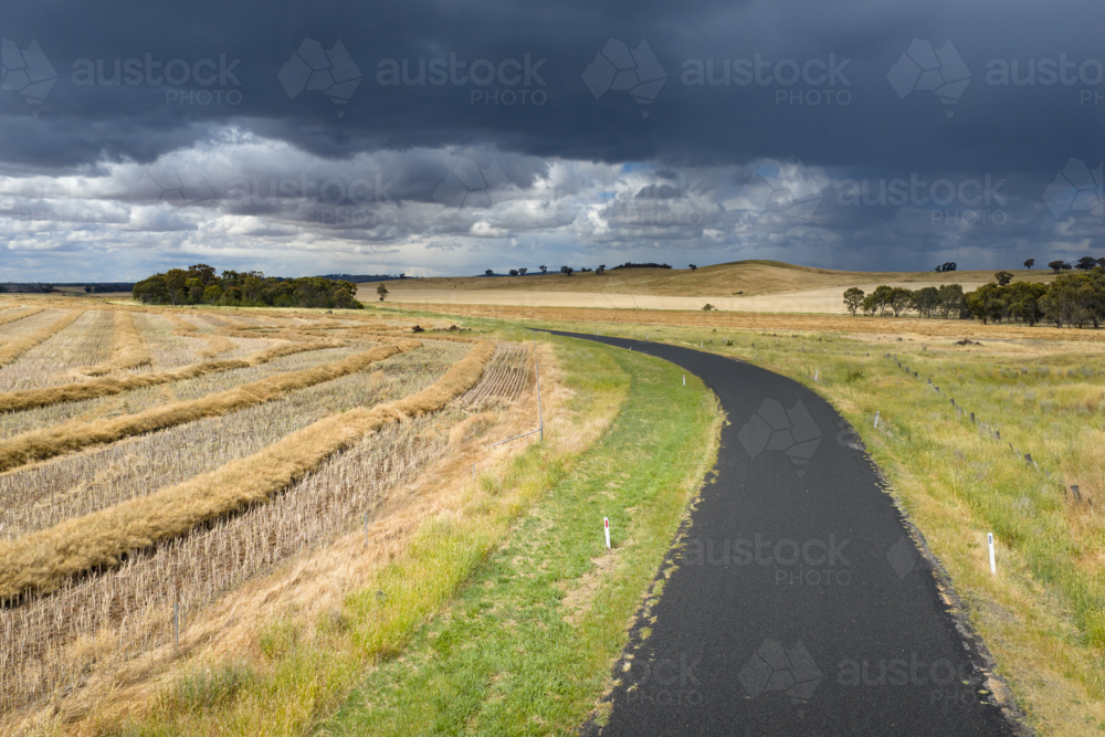 Aerial view of dark clouds and rain over a bend in a country road running through harvested farmland - Australian Stock Image