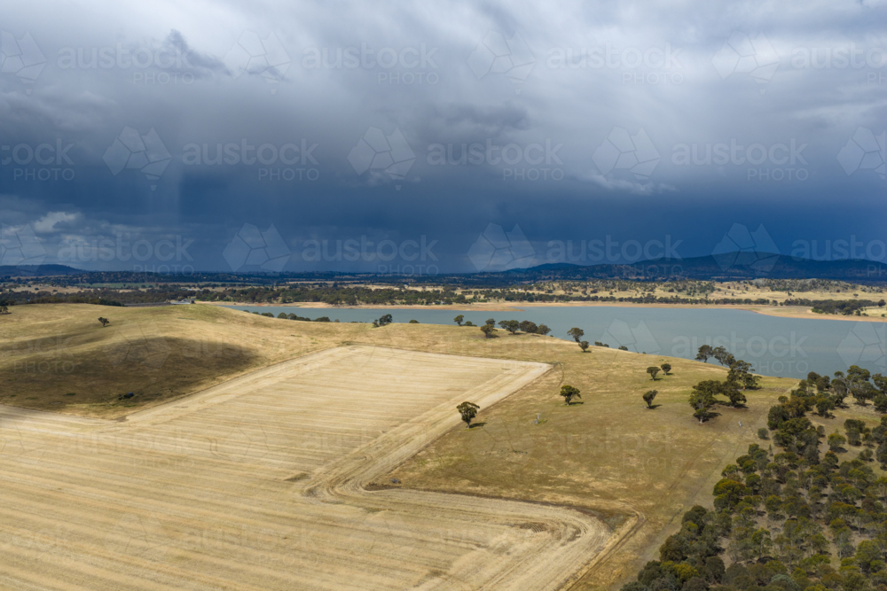 Aerial view of dry harvested farmland alongside a reservoir with rain falling from dark clouds above - Australian Stock Image