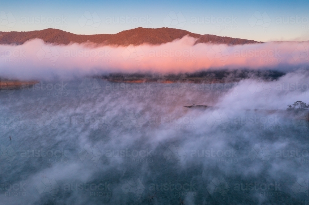 Aerial view of early morning light over a mountain lake covered with fog patches - Australian Stock Image