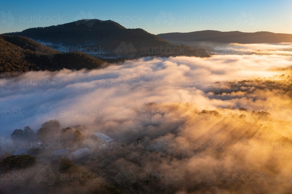 Aerial view of early morning sunshine on a mountain valley covered with fog - Australian Stock Image