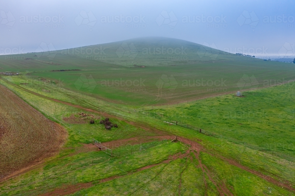 Aerial view of fog over green farmland and a low rounded hill - Australian Stock Image