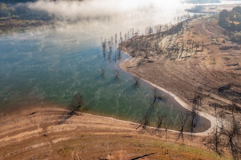 Aerial view of fog patches over a lake and dead trees along the shoreline near a small inlet - Australian Stock Image