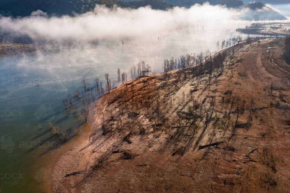 Aerial view of fog patches over a lake and dead trees casting long shadows along the shoreline - Australian Stock Image