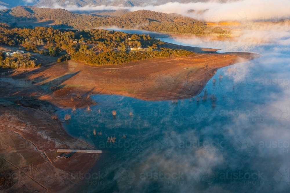 Aerial view of fog patches over a lake and morning sunshine over the shoreline - Australian Stock Image