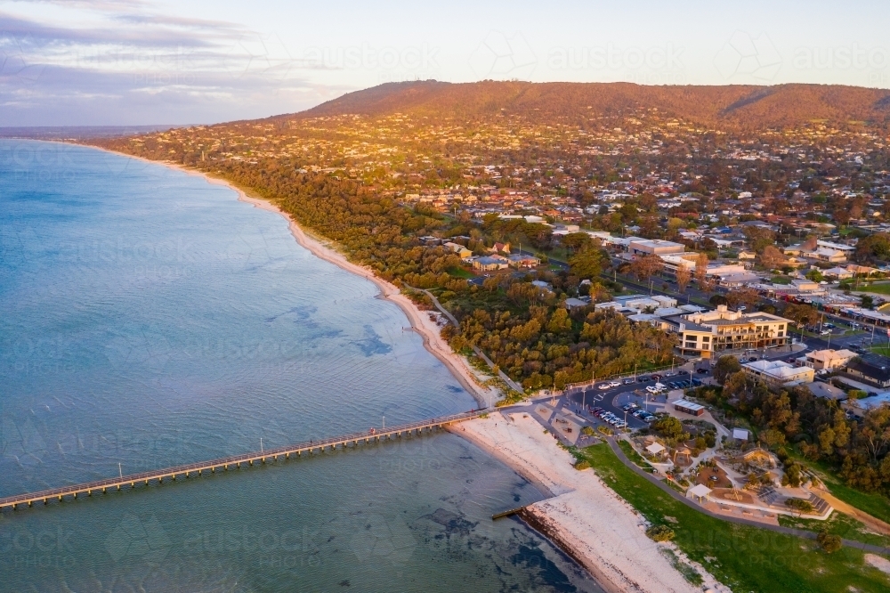 Aerial view of golden lighting over coastal reserve and jetty with mountains in background - Australian Stock Image
