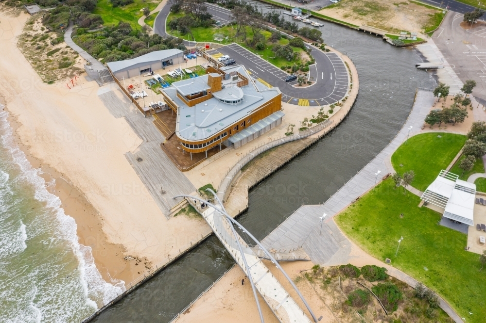 Aerial view of pedestrian bridge over a marina channel cutting through a beach, flowing out to sea - Australian Stock Image