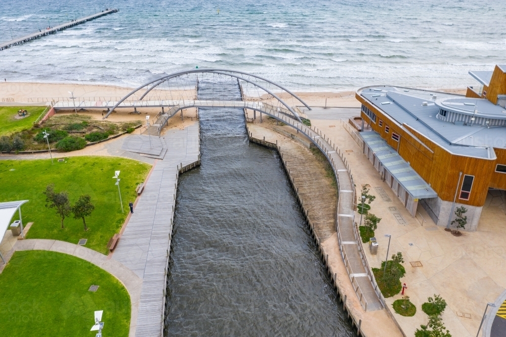 Aerial view of pedestrian bridge over a marina channel cutting through a beach, flowing out to sea - Australian Stock Image