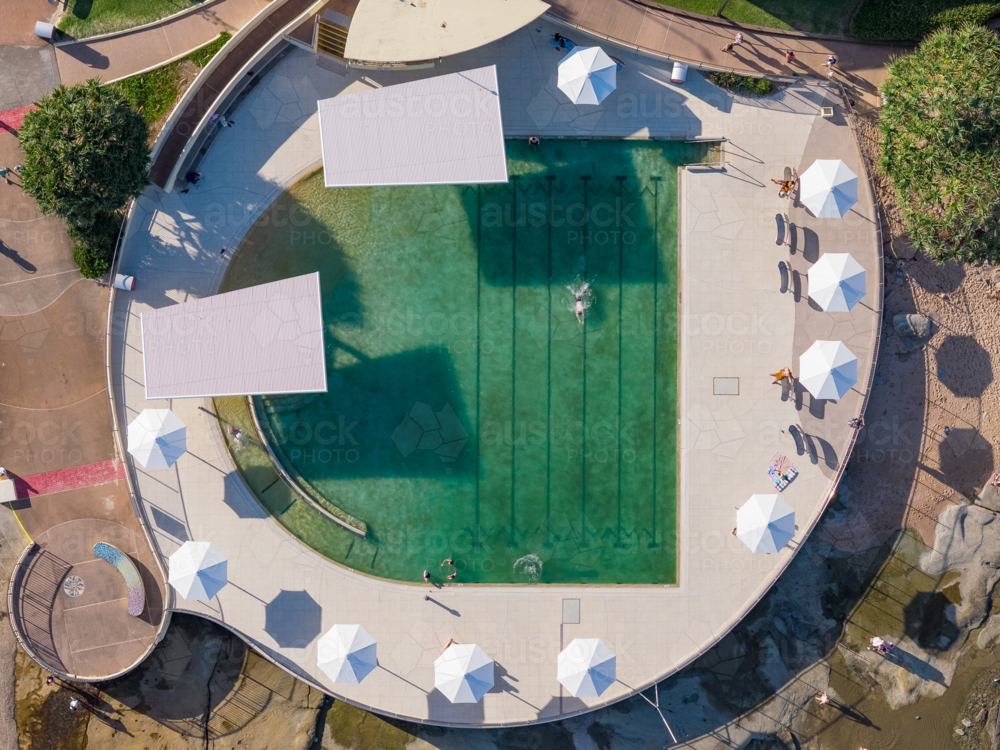 Aerial view of people swimming laps in a coastal pool with umbrellas alongside - Australian Stock Image