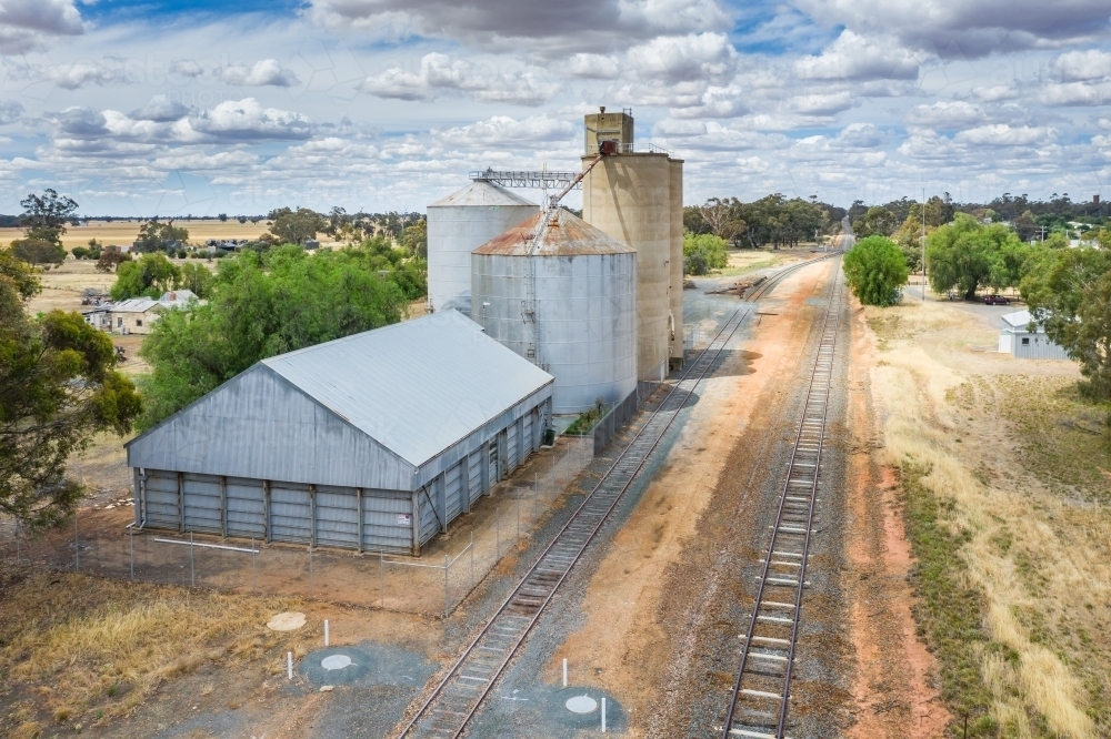 Aerial view of railway tracks running past a shed and grain silos in a rural town - Australian Stock Image