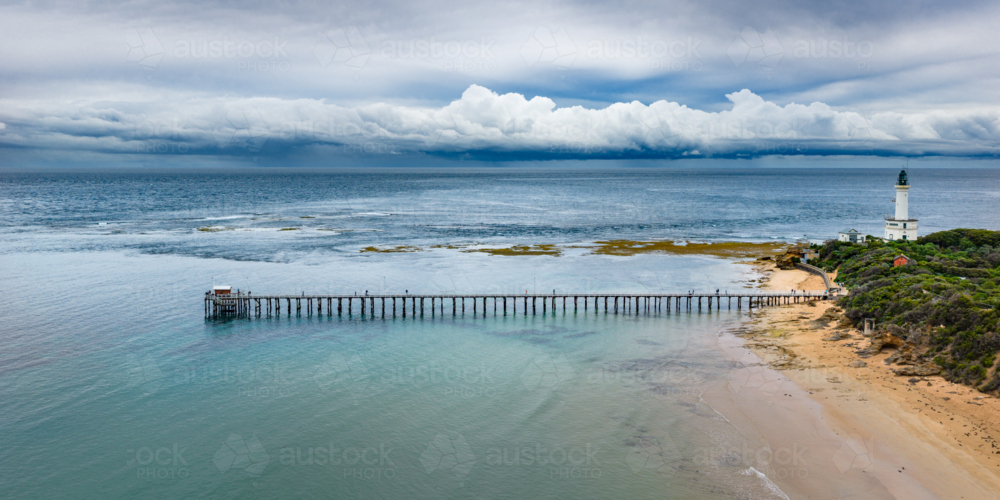 Aerial view of storm clouds approaching a coastal lighthouse and long narrow jetty over a calm bay - Australian Stock Image