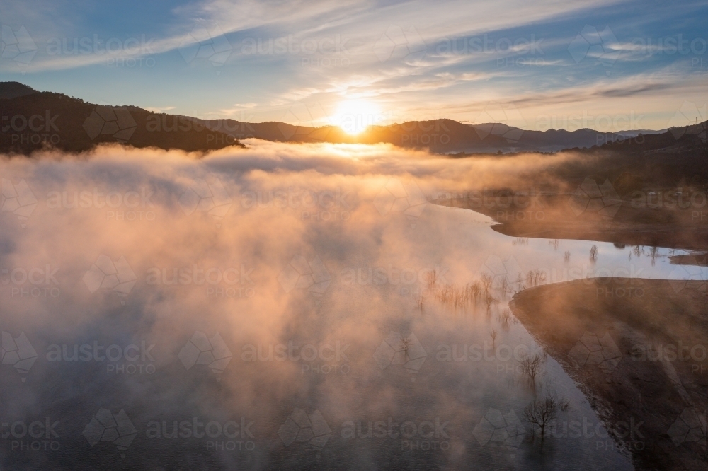 Aerial view of sunrise over mountains surrounding a lake covered in fog - Australian Stock Image
