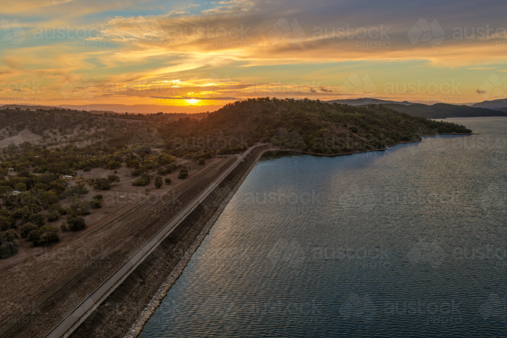 Aerial view of the Glenbawn Dam wall leading towards a sunset over the surrounding mountain range - Australian Stock Image